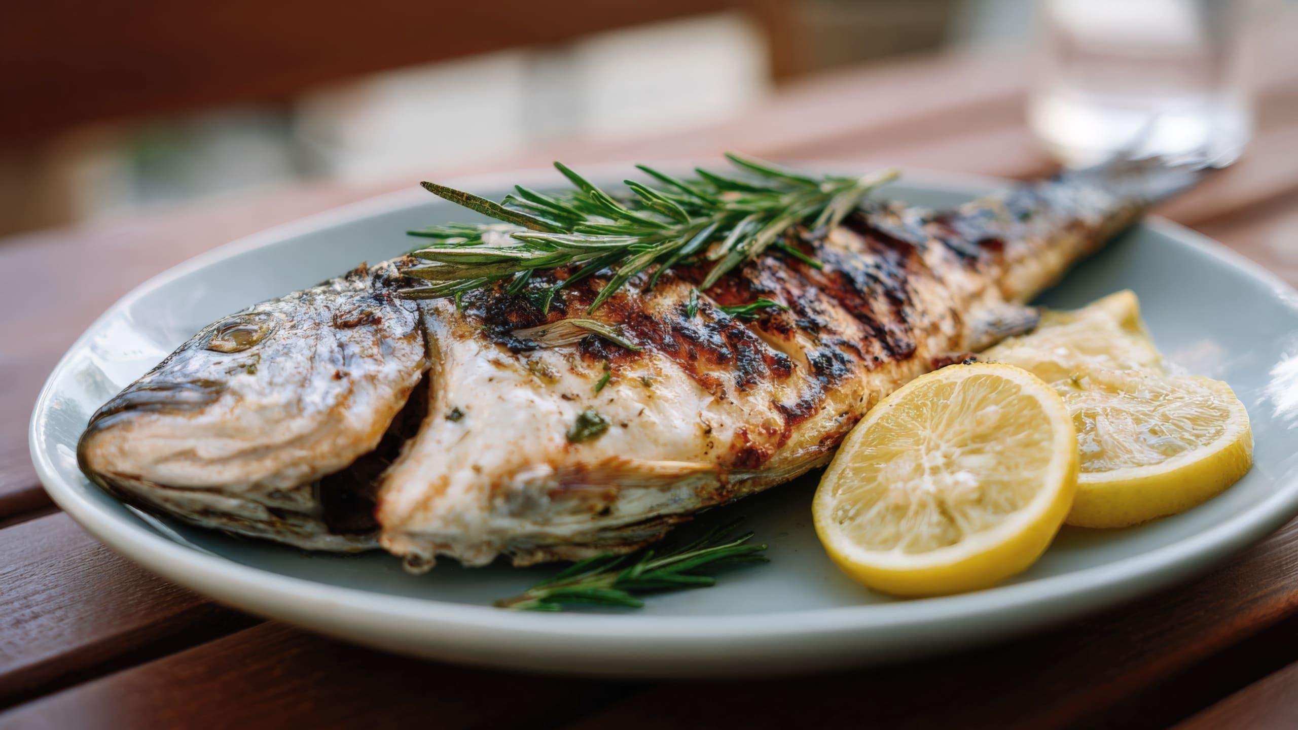 A whole grilled fish topped with fresh rosemary, served on a plate with lemon slices.