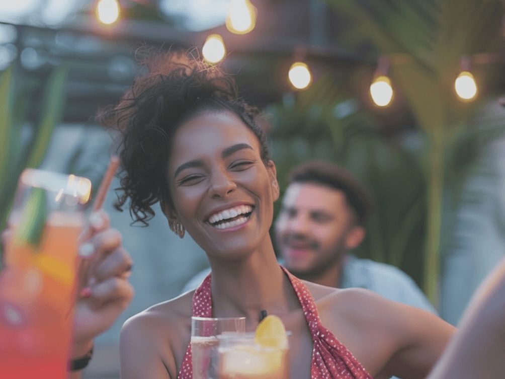 A woman laughing genuinely with friends at an evening gathering under string lights, drinks in hand.