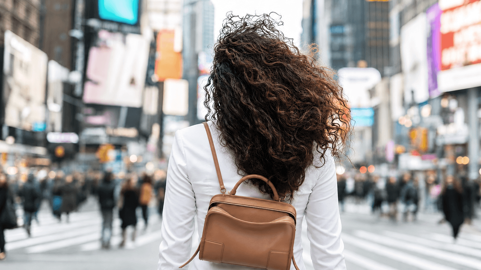 A woman with curly hair seen from behind, standing amid the illuminated billboards and crowds of a city square.