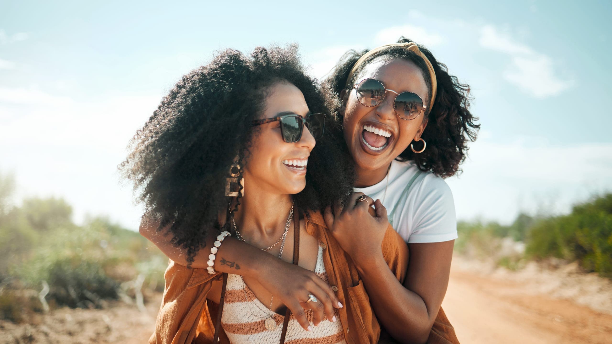 Two women laughing together outdoors on a dirt path, one with her arm around the other's shoulders, both wearing sunglasses in warm afternoon light.