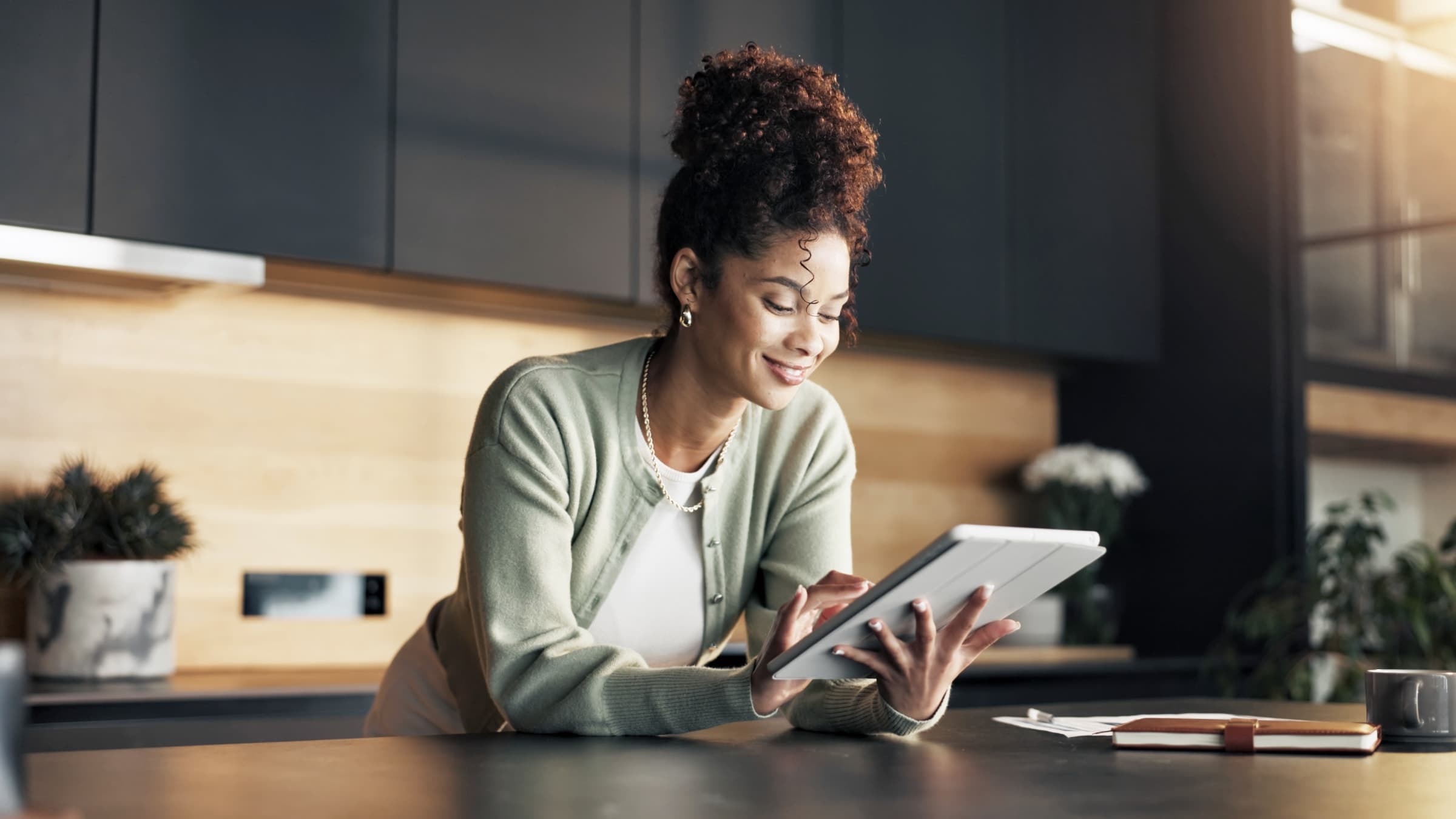 Woman in a sage cardigan smiling at a tablet while leaning on a kitchen counter, with a notebook and papers nearby.