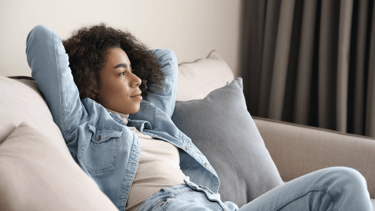 A woman relaxing on a couch in casual denim, hands behind her head, gazing out a window at ease.