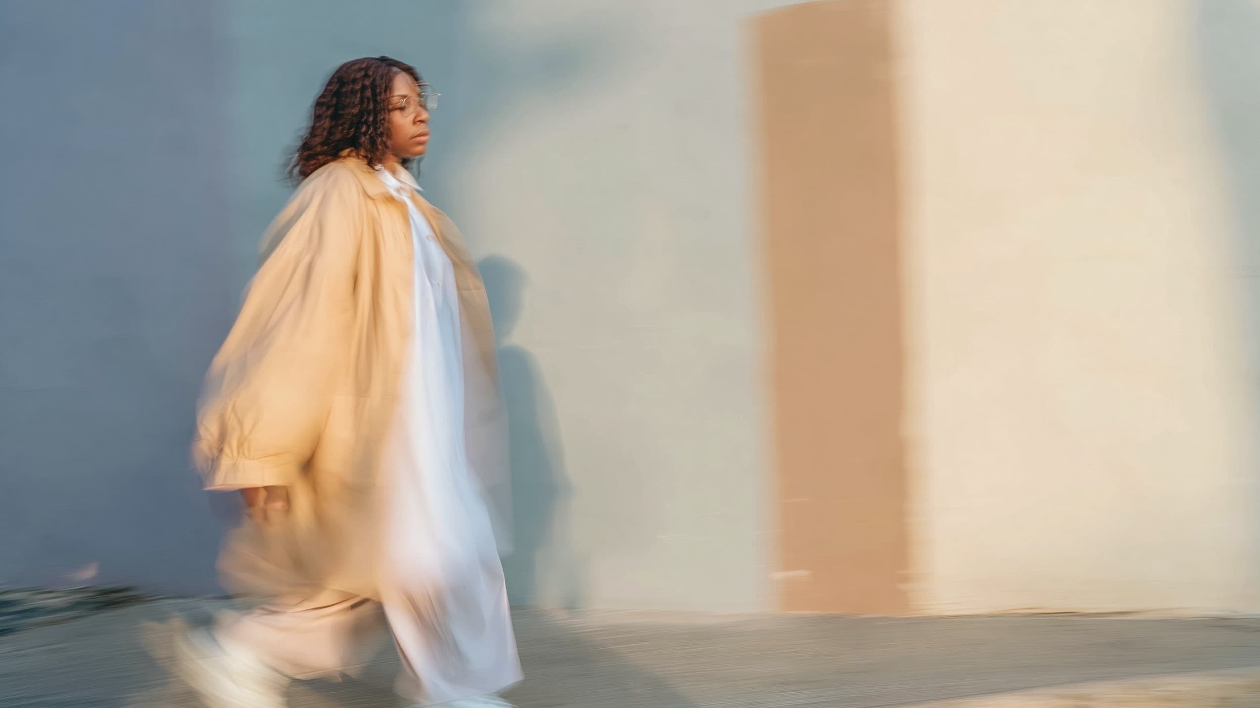 A woman in a camel coat walks forward in motion blur past a sunlit wall, glasses on, gaze ahead.