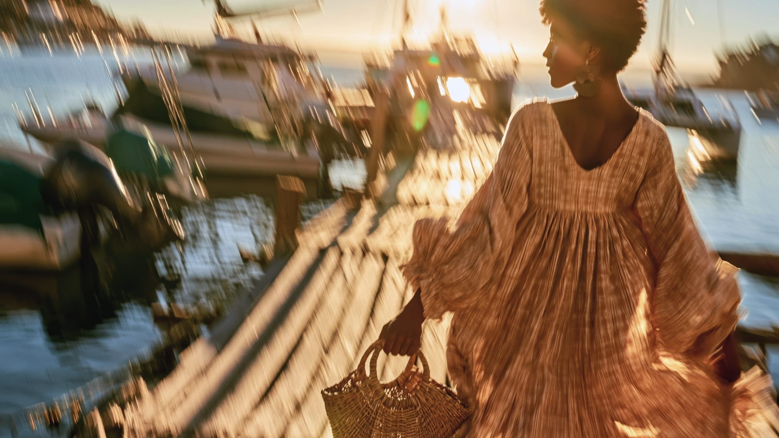 A woman in a flowing patterned dress walks along a sunlit dock carrying a woven basket, boats blurred in motion behind her.