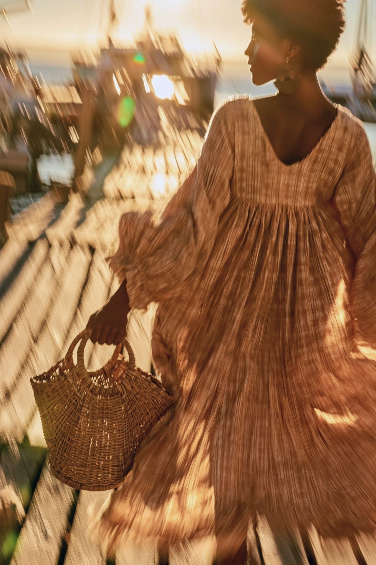 A woman in a flowing patterned dress walks along a sunlit dock carrying a woven basket, boats blurred in motion behind her.