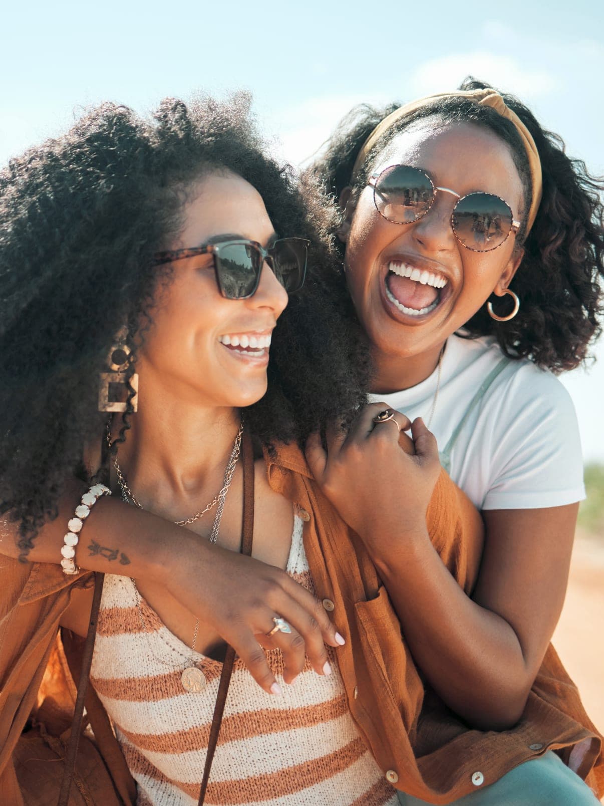 Two women laughing together outdoors on a dirt path, one with her arm around the other's shoulders, both wearing sunglasses in warm afternoon light.