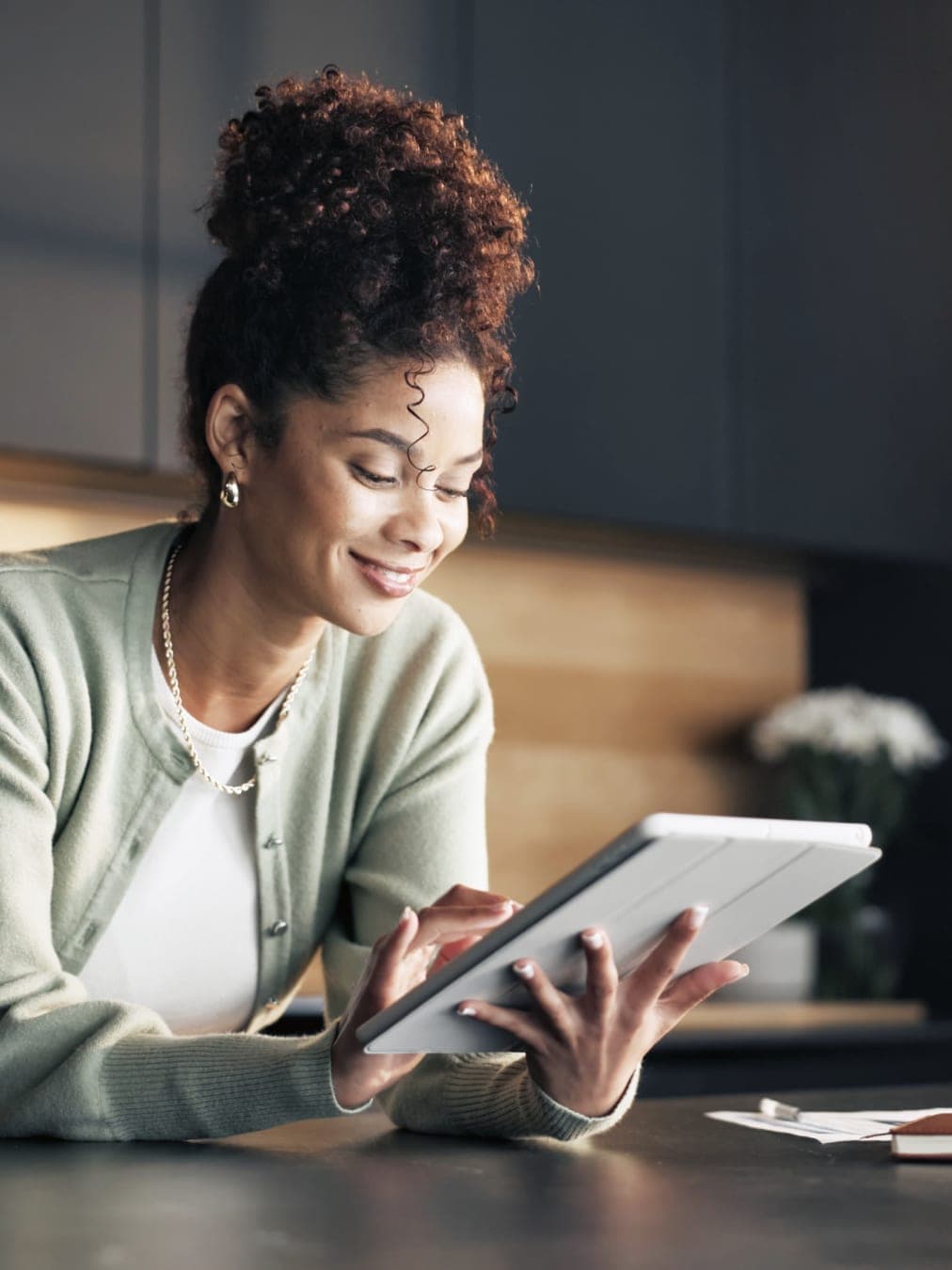 Woman in a sage cardigan smiling at a tablet while leaning on a kitchen counter, with a notebook and papers nearby.