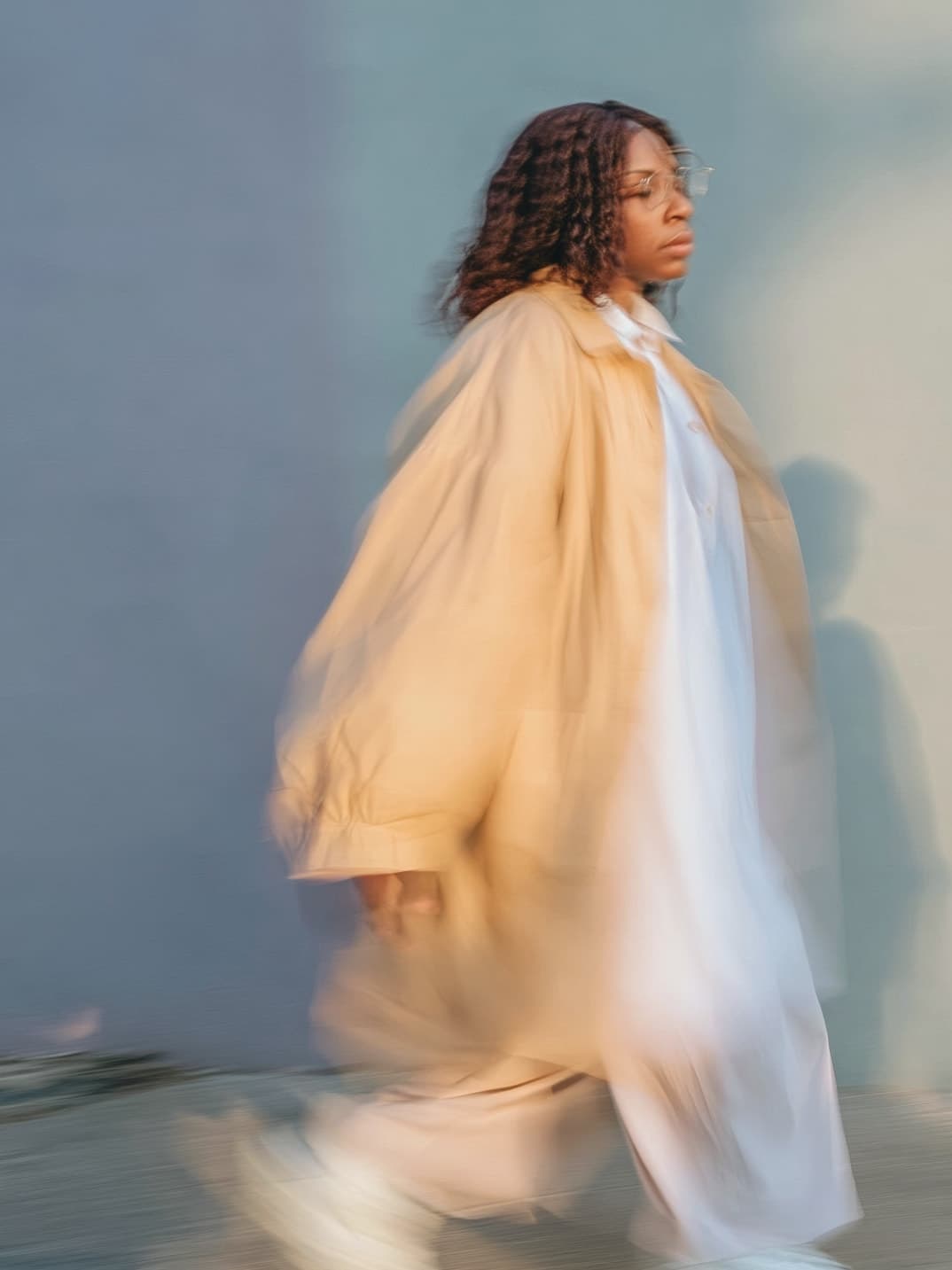 A woman in a camel coat walks forward in motion blur past a sunlit wall, glasses on, gaze ahead.