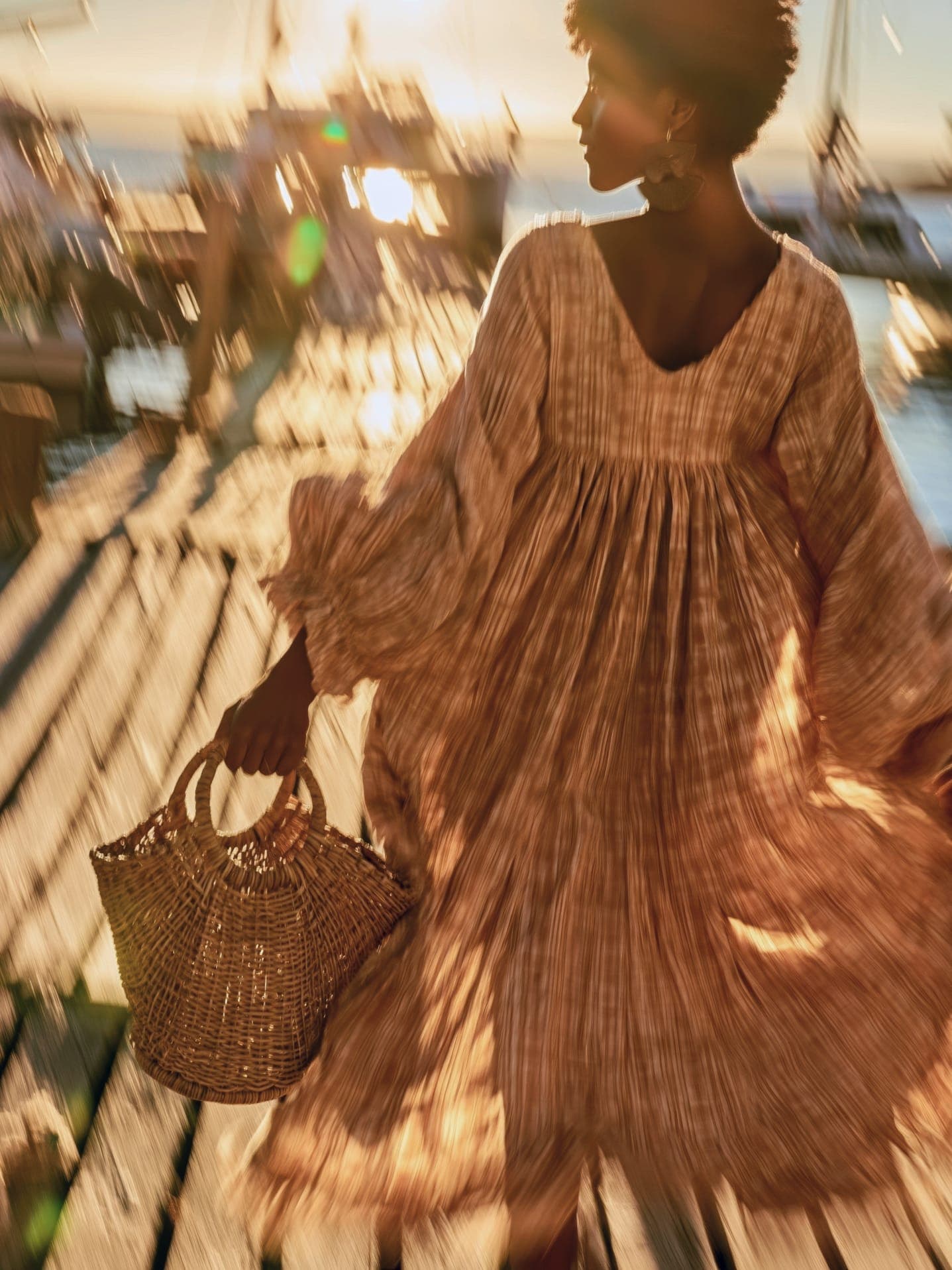 A woman in a flowing patterned dress walks along a sunlit dock carrying a woven basket, boats blurred in motion behind her.