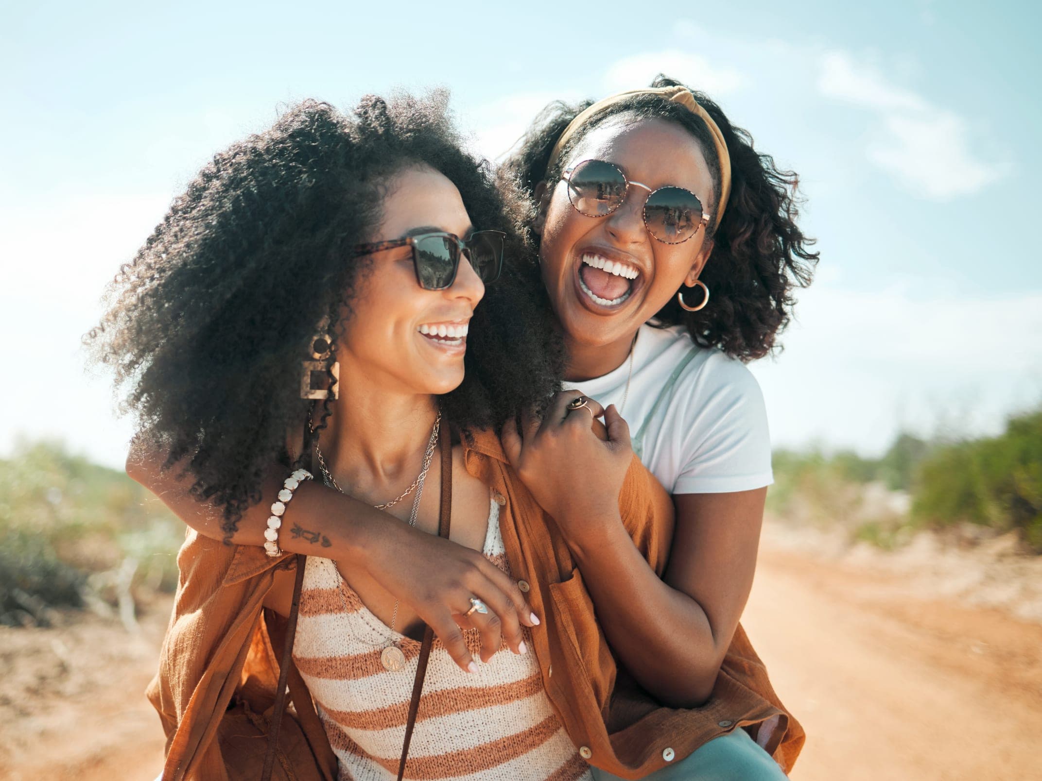 Two women laughing together outdoors on a dirt path, one with her arm around the other's shoulders, both wearing sunglasses in warm afternoon light.