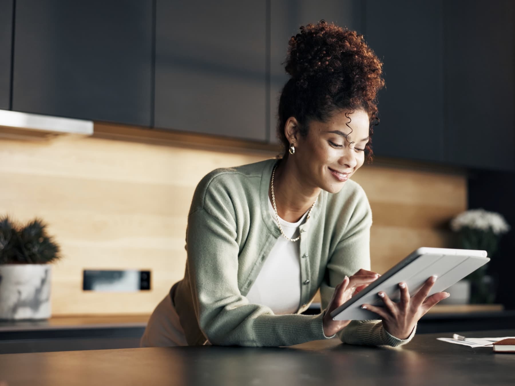 Woman in a sage cardigan smiling at a tablet while leaning on a kitchen counter, with a notebook and papers nearby.