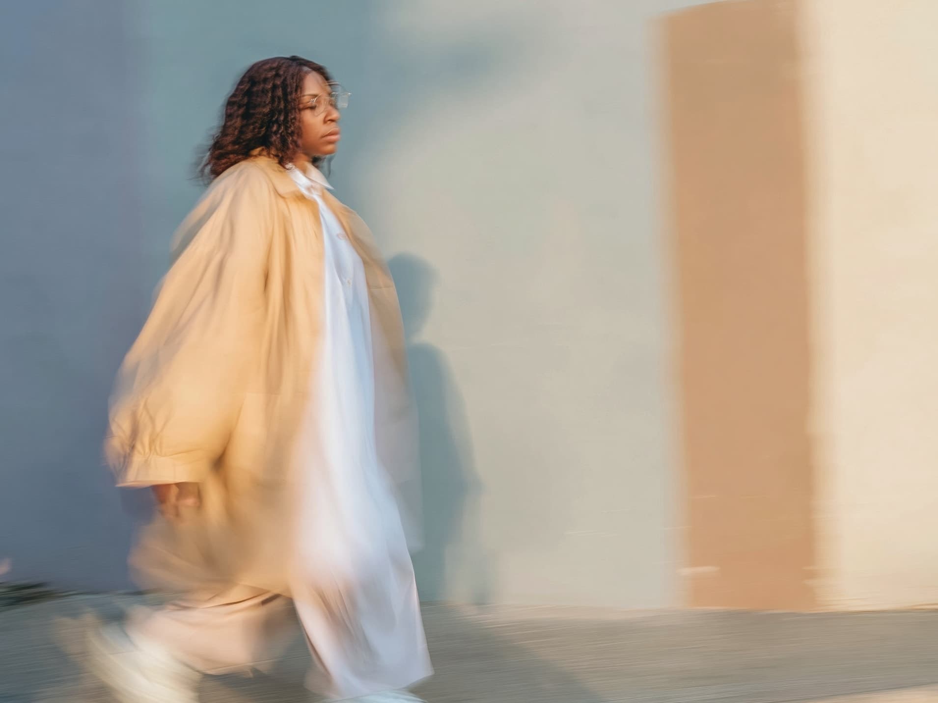 A woman in a camel coat walks forward in motion blur past a sunlit wall, glasses on, gaze ahead.