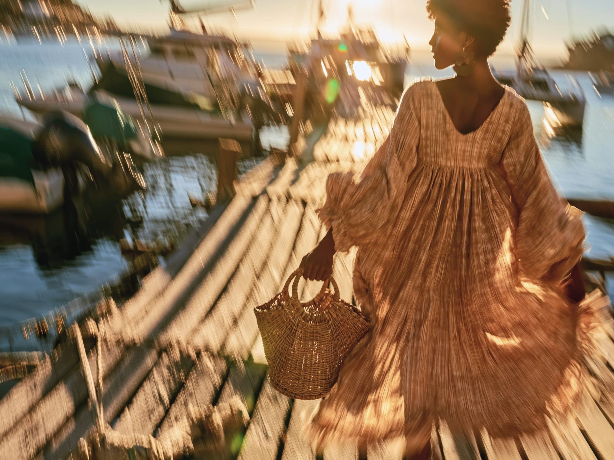 A woman in a flowing patterned dress walks along a sunlit dock carrying a woven basket, boats blurred in motion behind her.